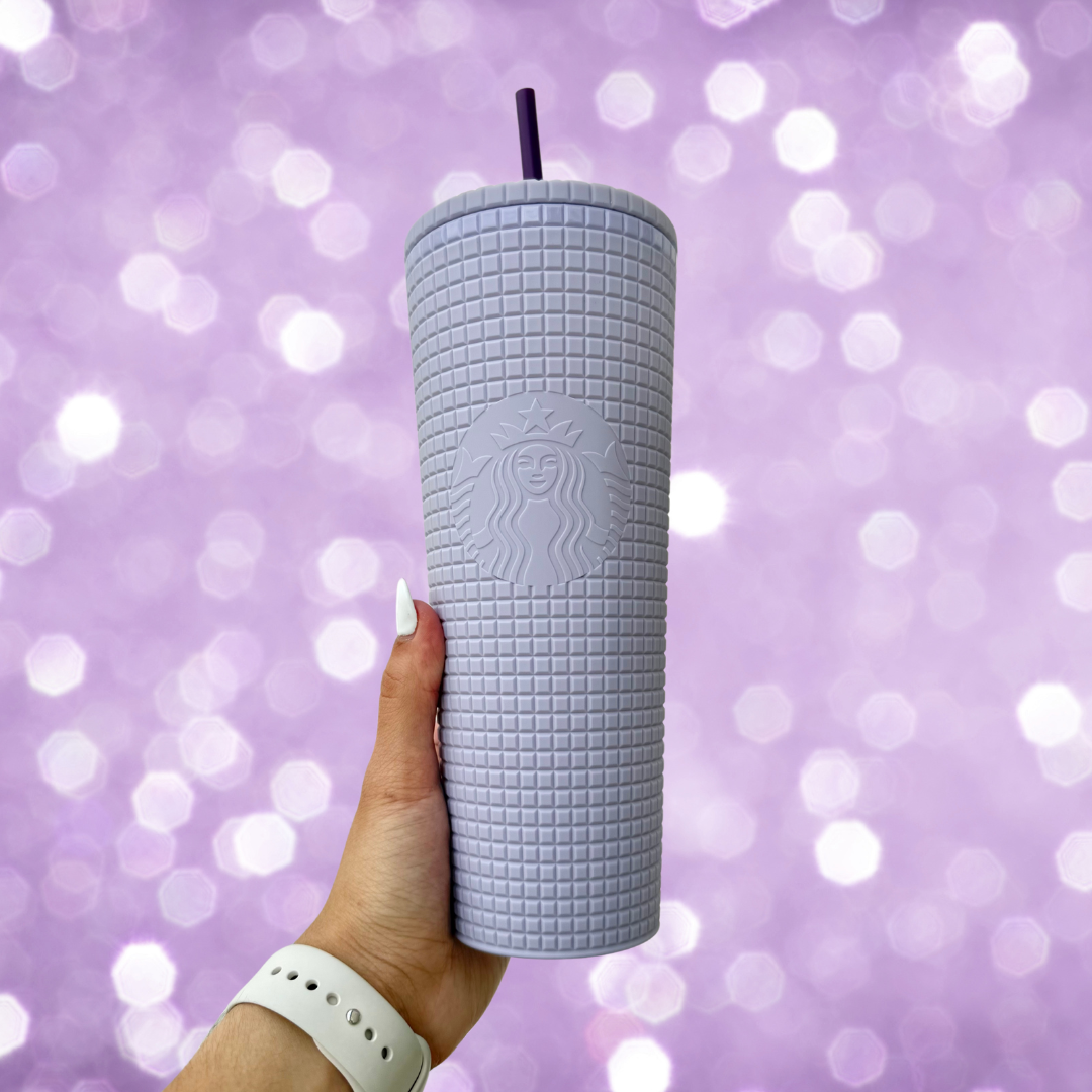 A hand holding a purple grid soft touch venti tumbler with a straw and twist-on lid against a purple sparkle background.
