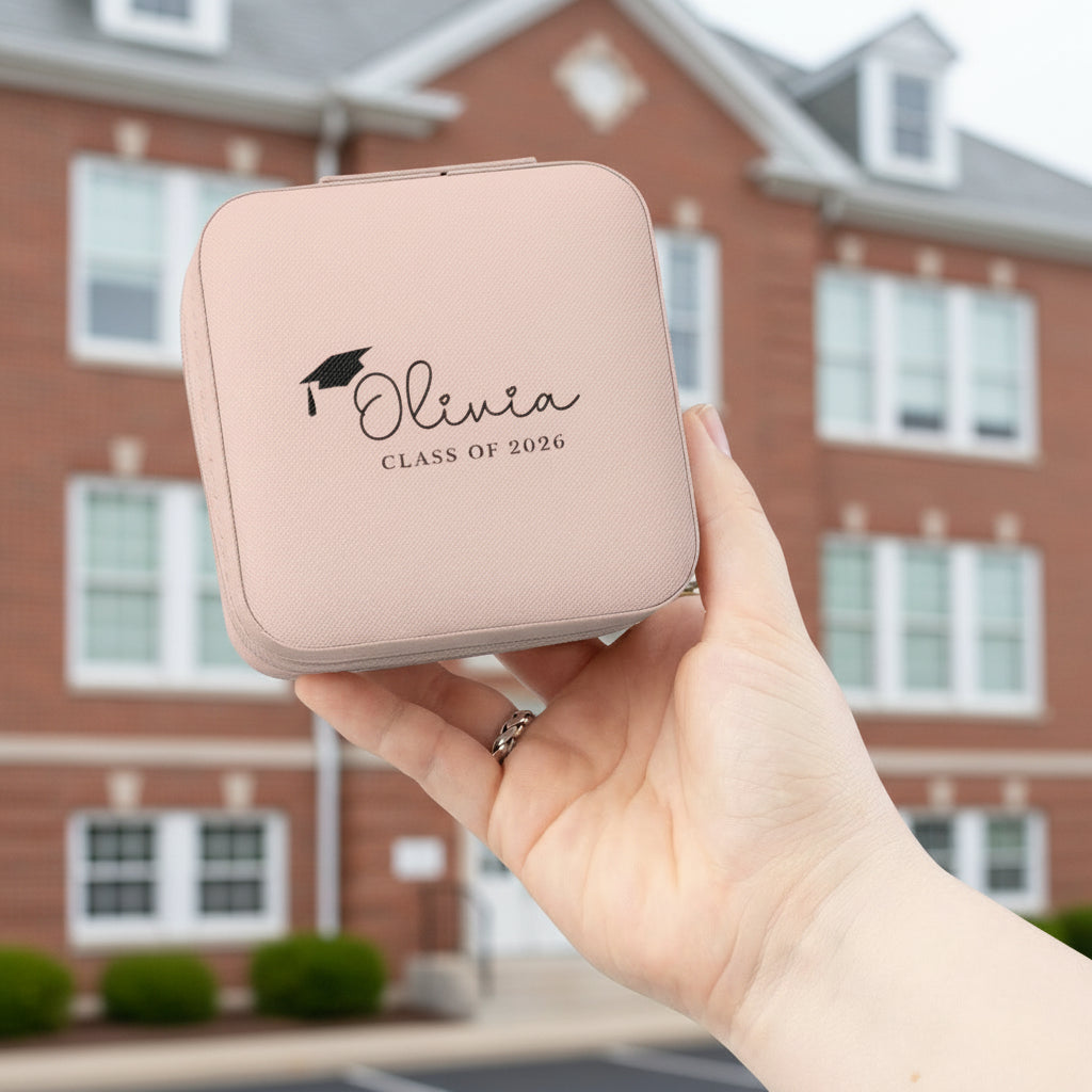 Pink jewelry case with 'Olivia Class of 2026' text held in a hand in front of a a school building