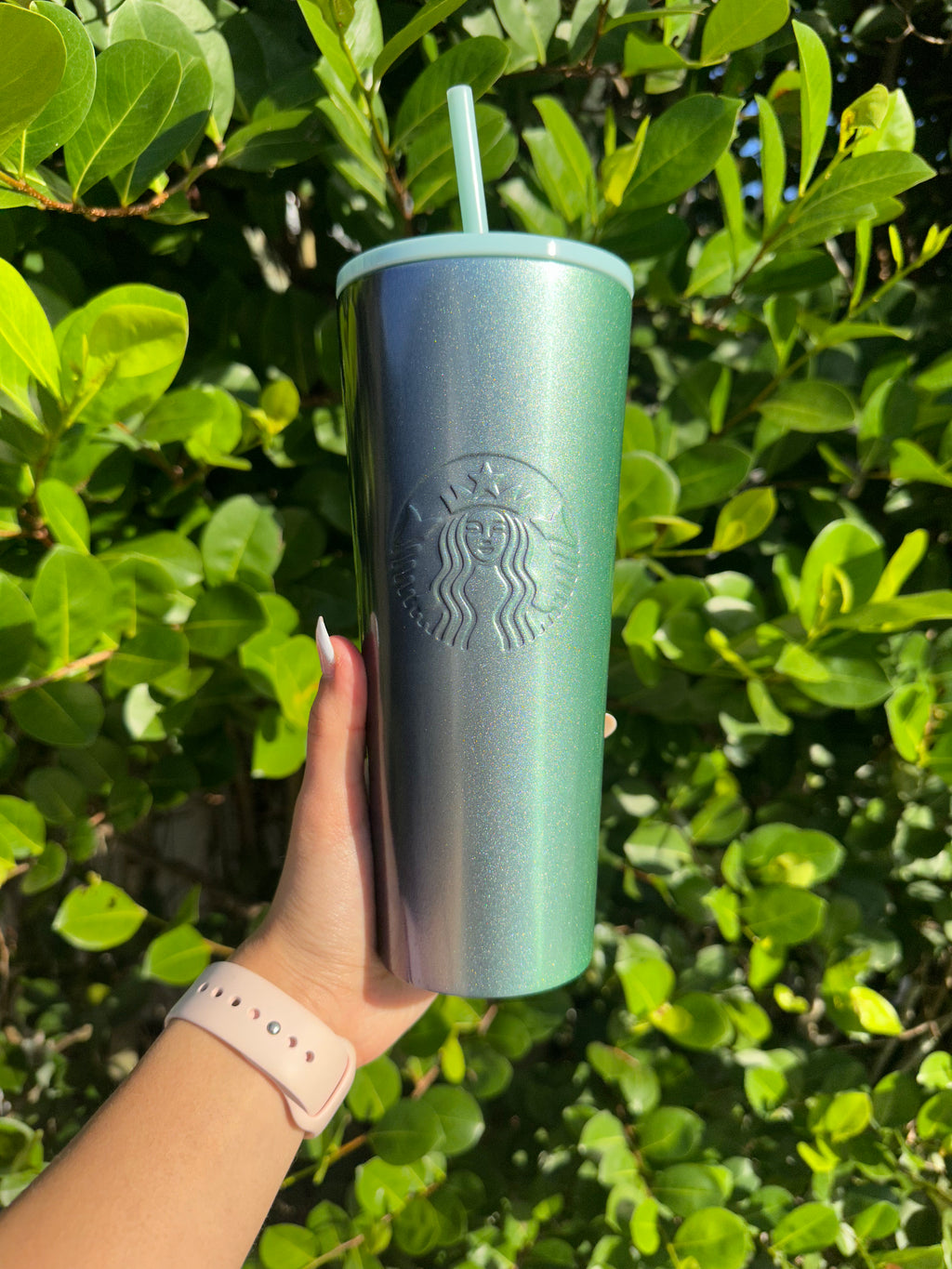 A hand holding a stainless steel tumbler with purple and green glitter ombre design, and the logo visible.